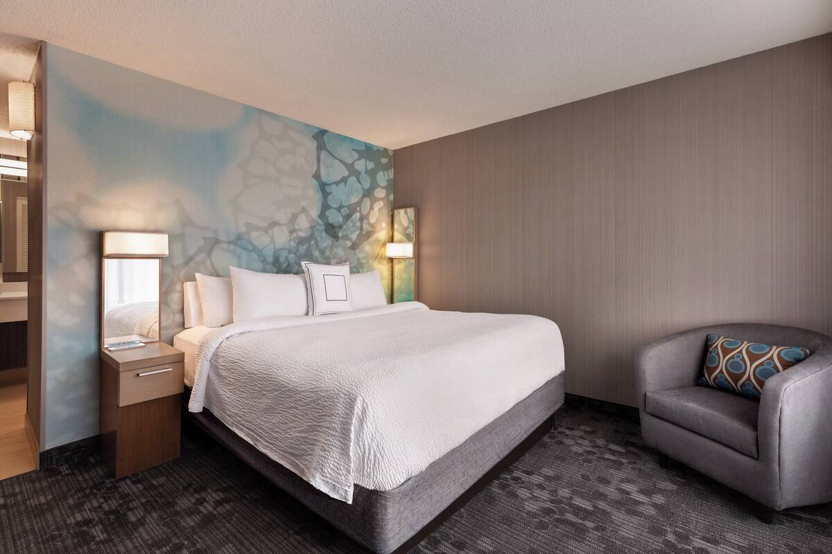 A hotel room with a large bed, a nightstand, a desk, a chair, and a wall with a textured mural.