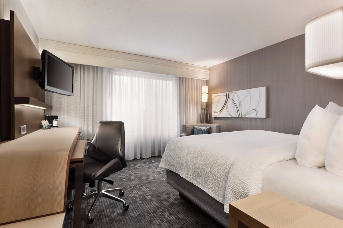 A hotel room with a large bed, a desk with a chair, a television mounted on the wall, and a window with curtains.