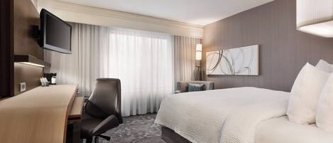 Habitación de hotel con una cama grande, un escritorio con silla, televisor montado en la pared y una ventana con cortinas.
