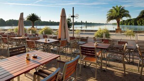 An outdoor dining area with tables and chairs, a palm tree, and a view of a lake. - Courtyard by Marriott Hannover Maschsee (Hannover)