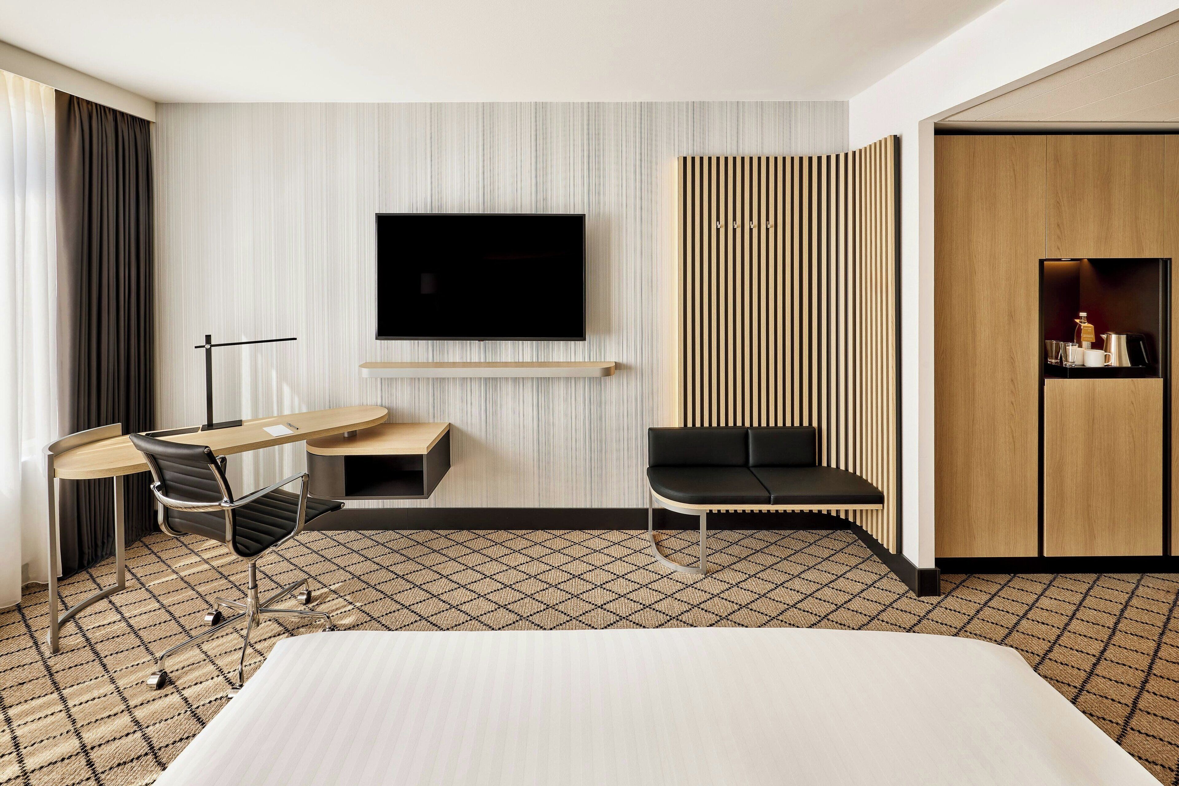 A modern hotel room with a large bed, a desk with a chair, a flat-screen TV mounted on the wall, a wooden wardrobe, and a patterned floor.