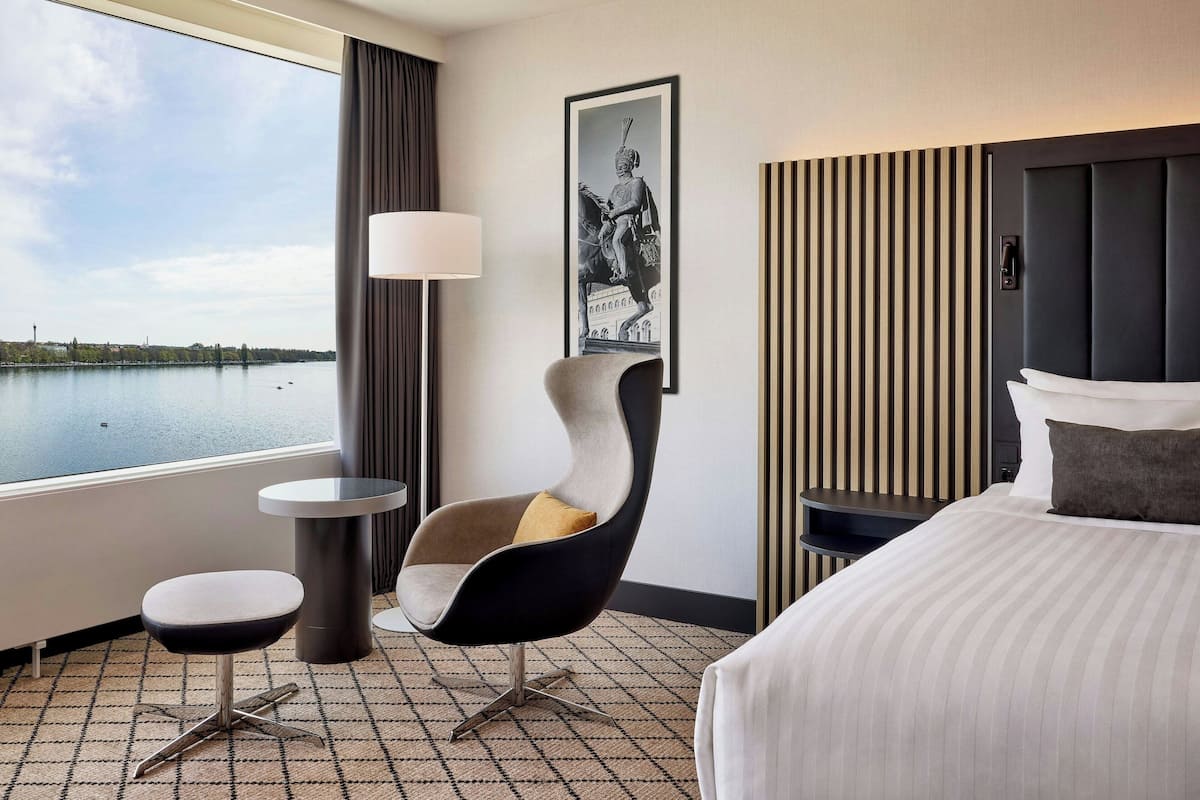 A hotel room with a bed, a modern chair, a small table, a lamp, and a framed picture on the wall.