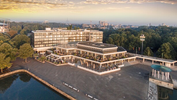 A multi-story hotel with a courtyard and outdoor seating area, surrounded by trees and a body of water. - Courtyard by Marriott Hannover Maschsee (Hannover)