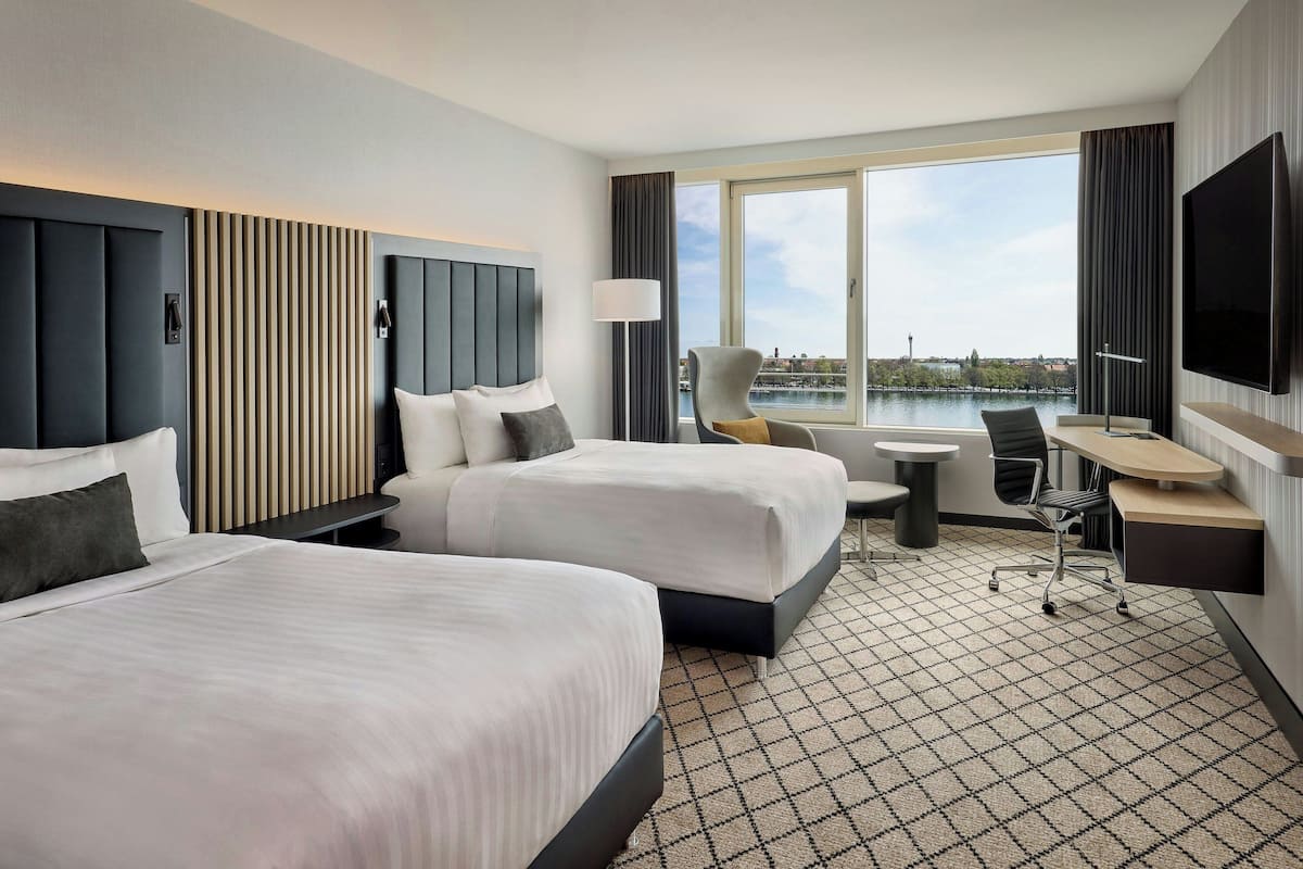 A hotel room with two beds, a desk, a chair, a TV, and a large window with a view of a body of water.