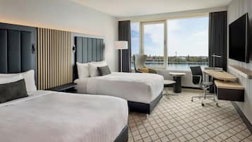 Ein Hotelzimmer mit zwei Betten, einem Schreibtisch, einem Sessel, einem Fernseher und einem großen Fenster mit Blick auf ein Gewässer.