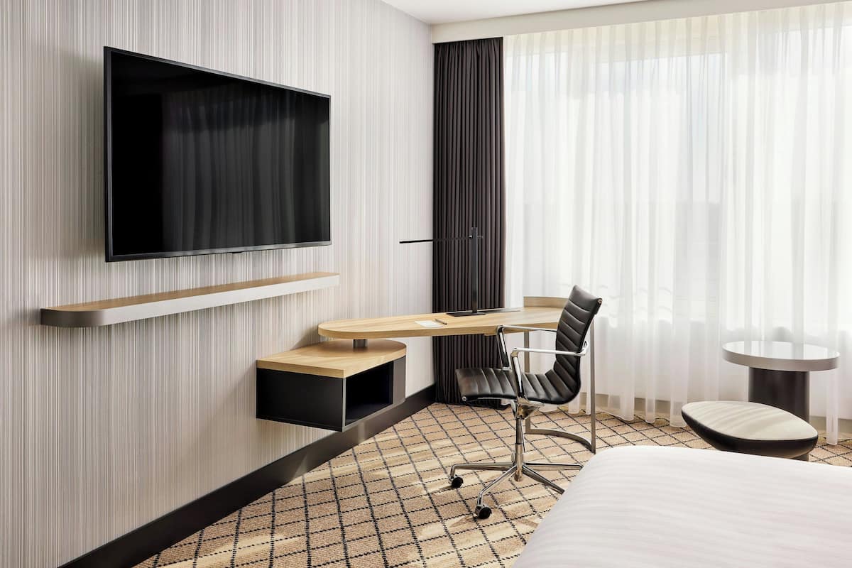 A modern hotel room with a flat-screen TV mounted on the wall, a desk with a chair, a small round table, and a bed with a patterned rug.