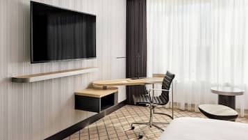Habitación de hotel moderna con una televisión de pantalla plana montada en la pared, un escritorio con una silla, una mesita redonda pequeña y una cama con un tapete estampado.