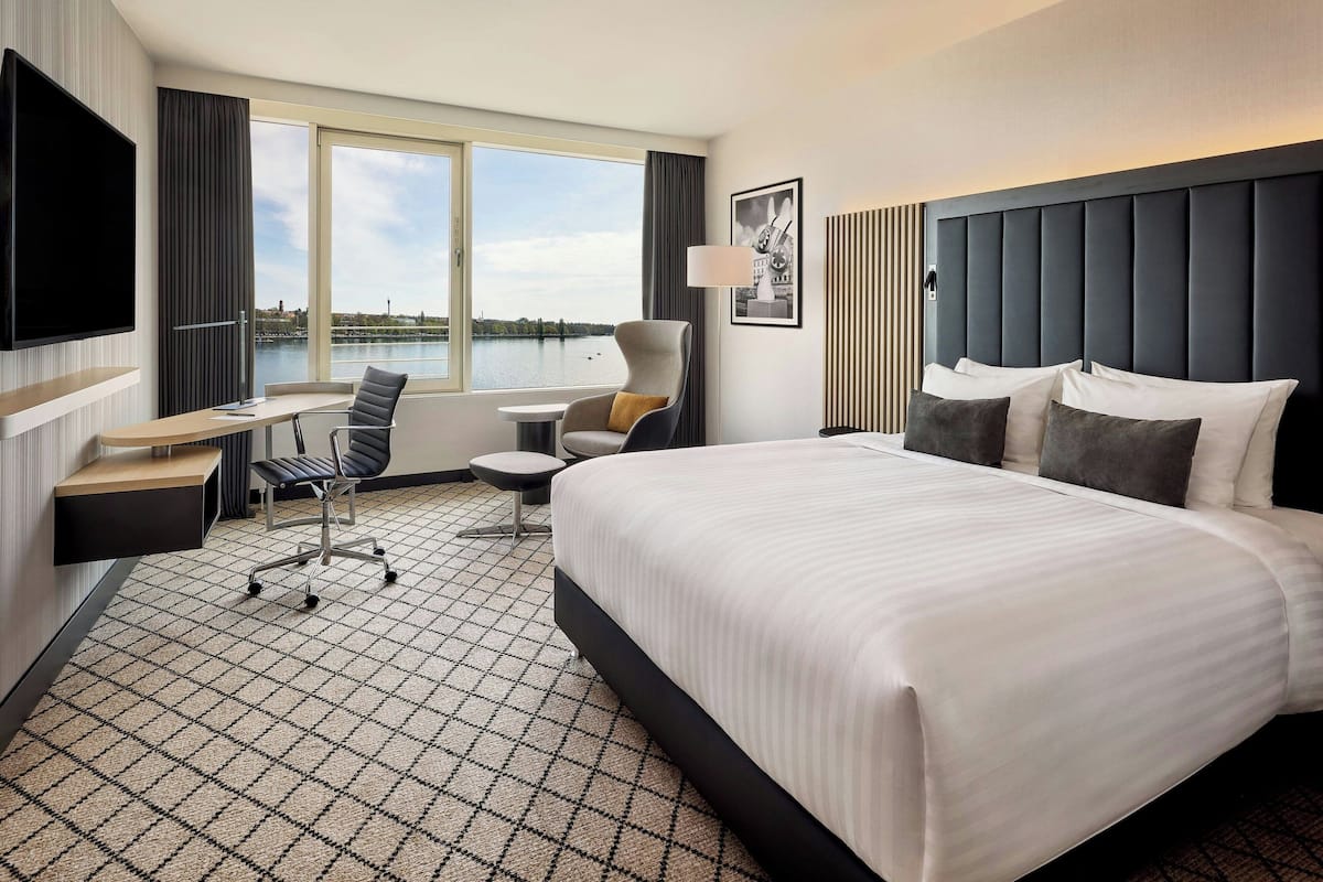 A hotel room with a large bed, a desk, a chair, a TV, and a view of a body of water.