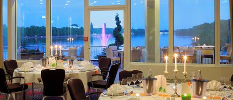Un restaurante con mesas dispuestas para comer, amplias ventanas con vista a un lago y una fuente, y velas en las mesas.