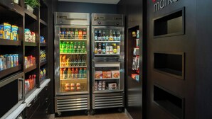 A compact, well-lit room with two large, stainless steel refrigerators stocked with beverages and snacks. Shelves on the left hold various packaged goods.