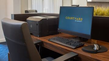 A hotel room with a wooden desk, a computer, a printer, and a chair.