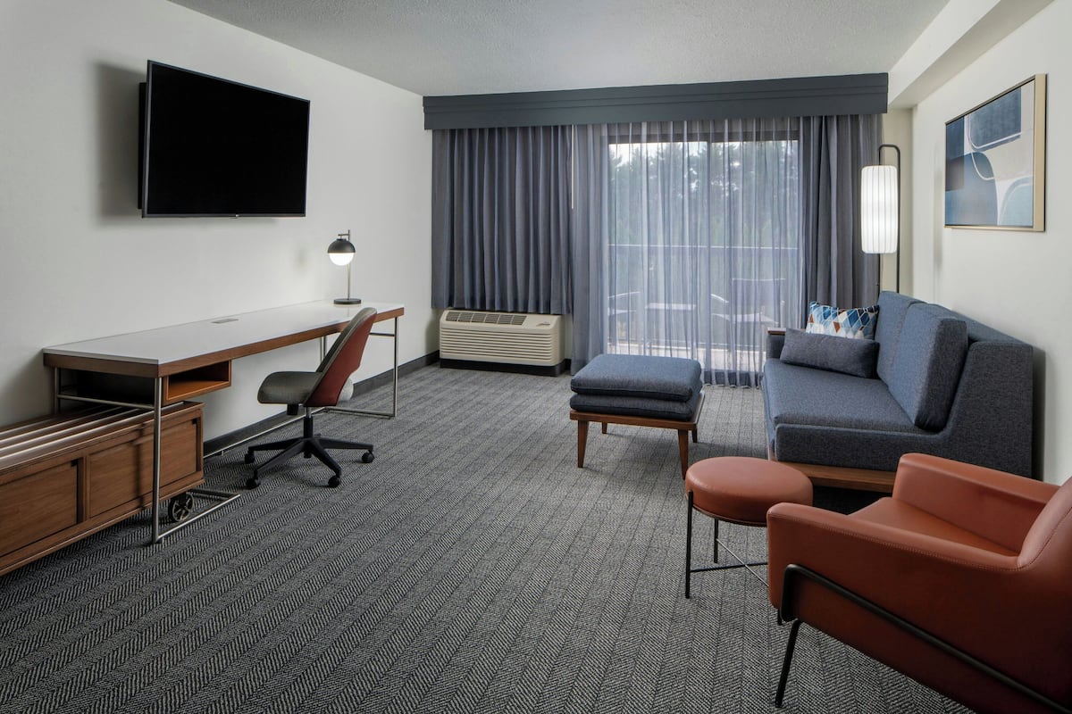 Quarto de hotel com televisão de tela plana, escrivaninha, cadeira, sofá, puf e uma grande janela com cortinas.