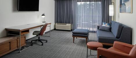 Ett hotellrum med en platt-TV, ett skrivbord, en stol, en soffa, en pall och ett stort fönster med gardiner.
