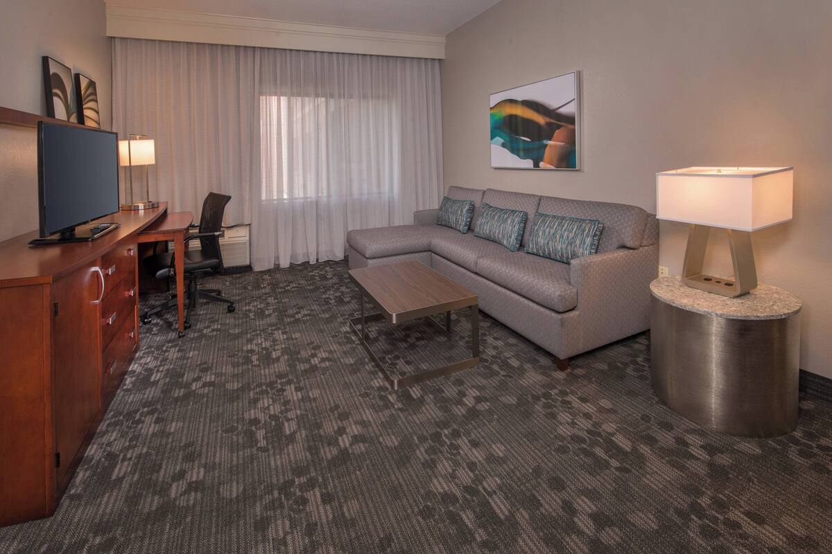 A hotel room with a grey sofa, a wooden desk with a TV, a lamp, and a painting on the wall.
