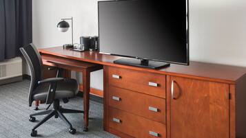 A hotel room with a wooden desk, a black office chair, a flat-screen TV, a coffee maker, and a lamp.