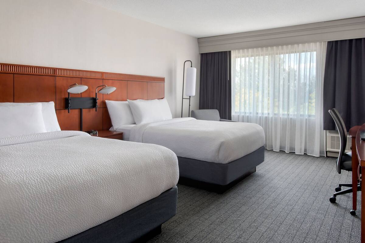 A hotel room with two beds, a desk, a chair, a lamp, and a large window with curtains.
