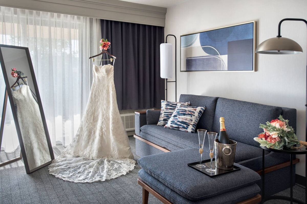 Een kamer met een grijze bank, een spiegel, een bruidsjurk, een champagnefles en een boeket.