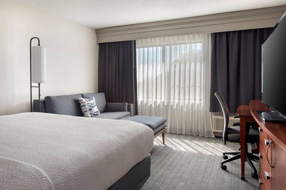 A hotel room with a large bed, a sofa, a desk with a flat-screen TV, a chair, and a window with curtains.