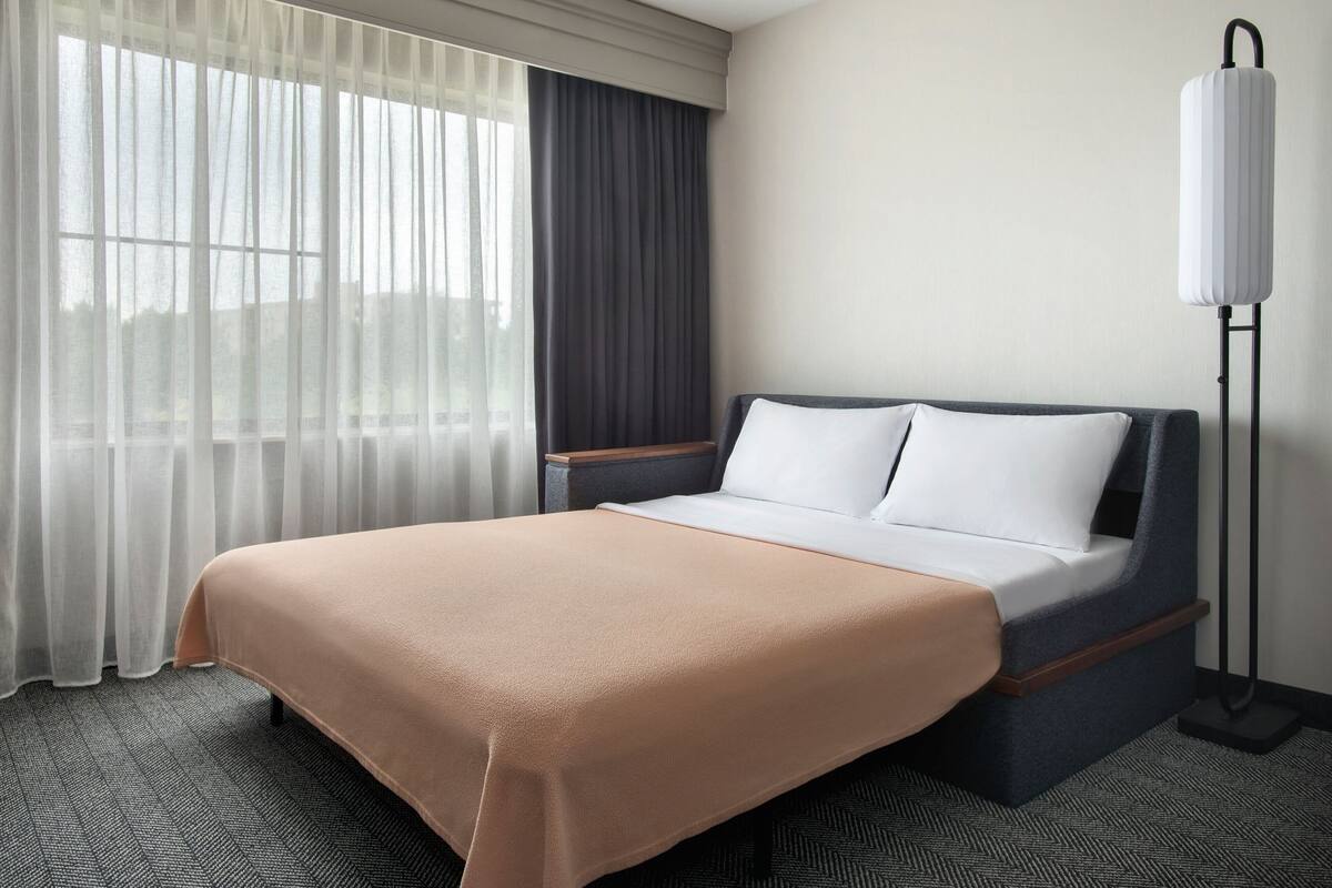 Een hotelkamer met een bed, nachtkastje, lamp en een raam met gordijnen.