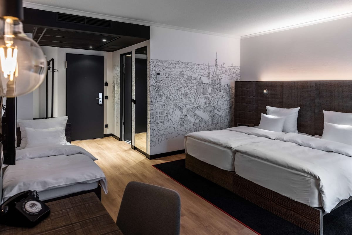 A hotel room with a large bed, a smaller bed, a desk, a chair, a lamp, and a wall mural.