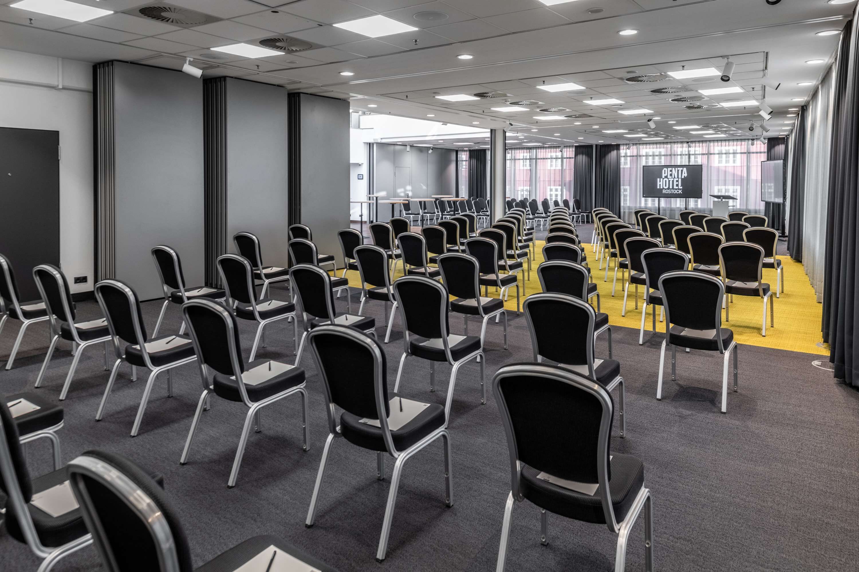Un salón de conferencias con hileras de sillas, un atril y una gran pantalla que muestra 'PENTA HOTEL ROSTOCK'.