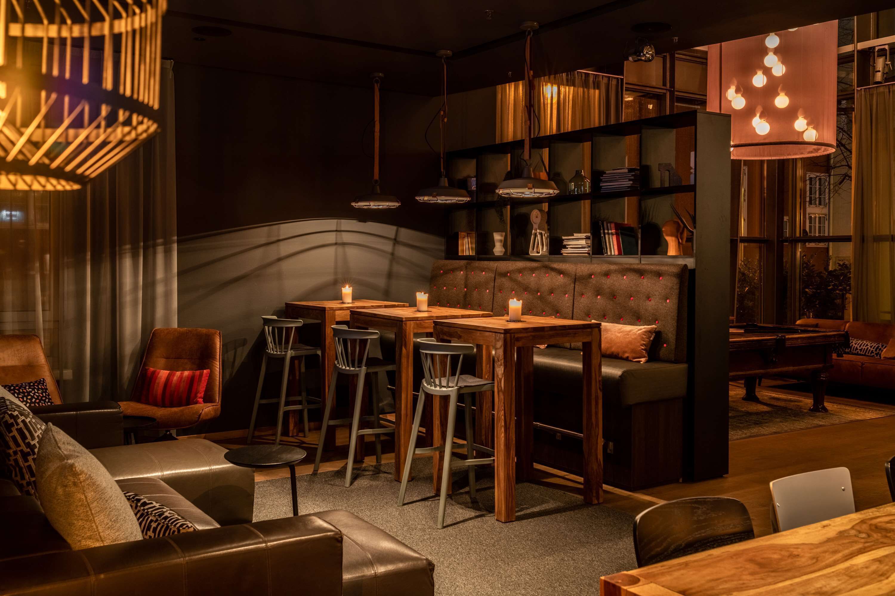 A cozy lounge area with leather sofas, wooden tables, and bar stools. There are bookshelves with books and decorative items, and a piano in the background.