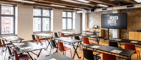 A conference room with tables and chairs, a large screen displaying 'PENTA HOTEL ROSTOCK', and large windows with a view of buildings.