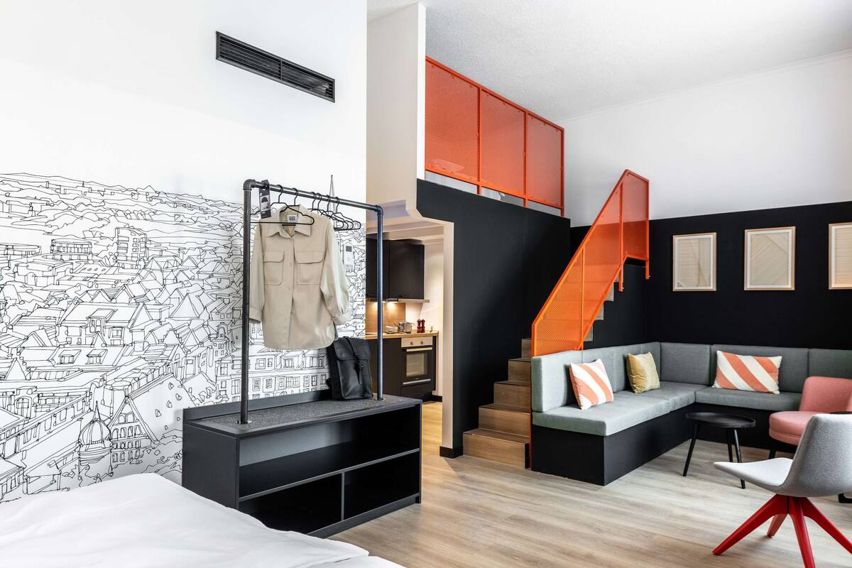 A modern hotel room with a black and white cityscape mural, a black console table, a gray sofa, a red chair, and a staircase with orange railings.