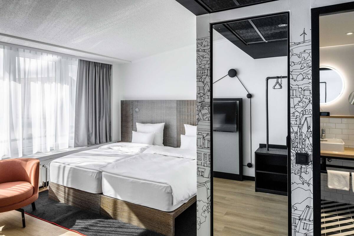 A modern hotel room with a large bed, a sitting area with a chair, a TV, and a bathroom with a mirror and sink.