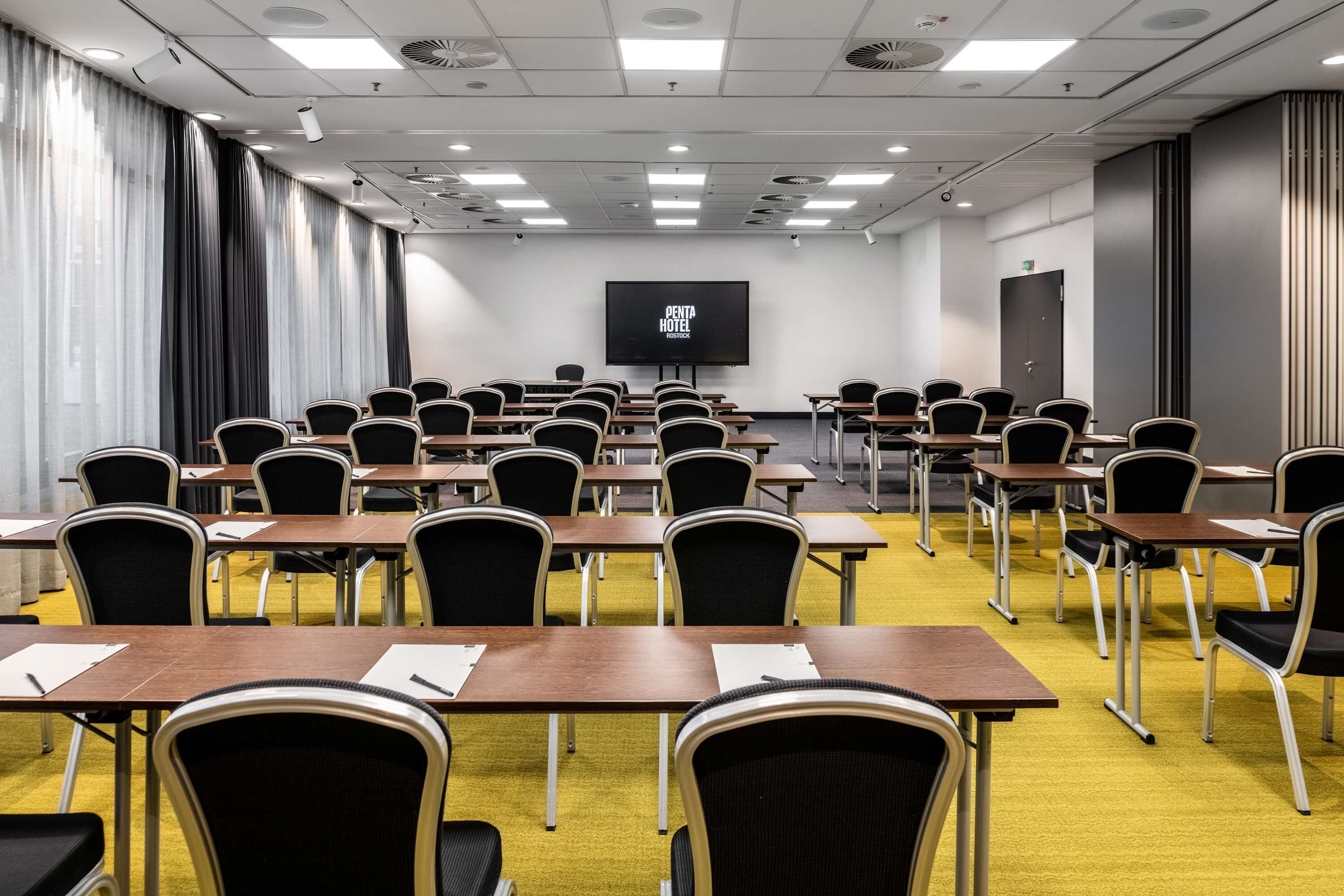 Ein Konferenzraum mit Reihen von Tischen und Stühlen, einem großen Bildschirm und einer Tür im Hintergrund.