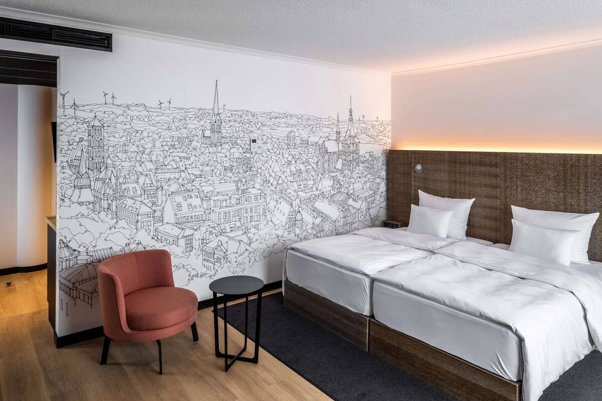 A hotel room with a large wall mural of a cityscape, a bed with white linens, a pink chair, and a small round table.