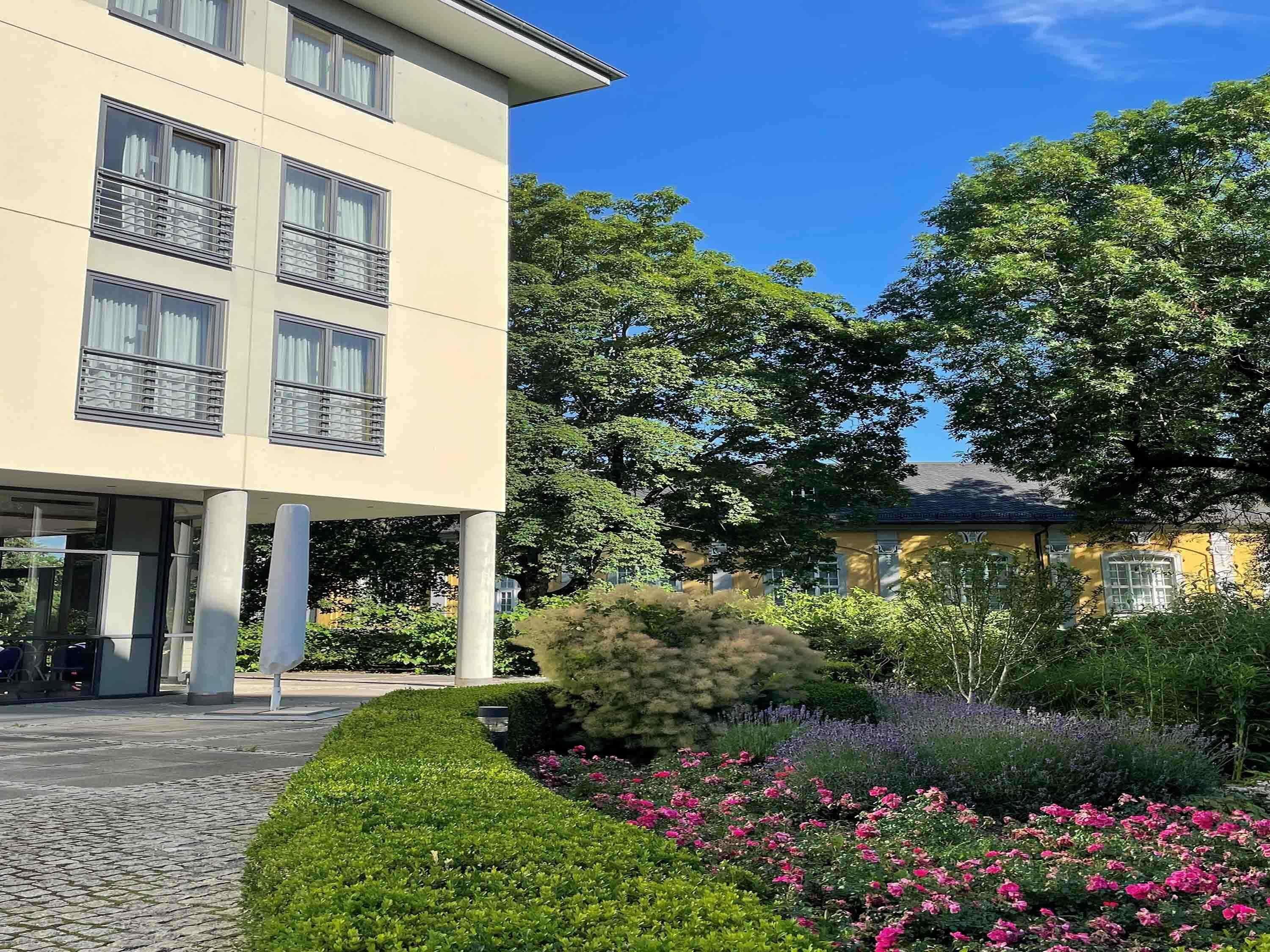 A modern building with balconies, a paved entrance, and a garden with pink flowers.