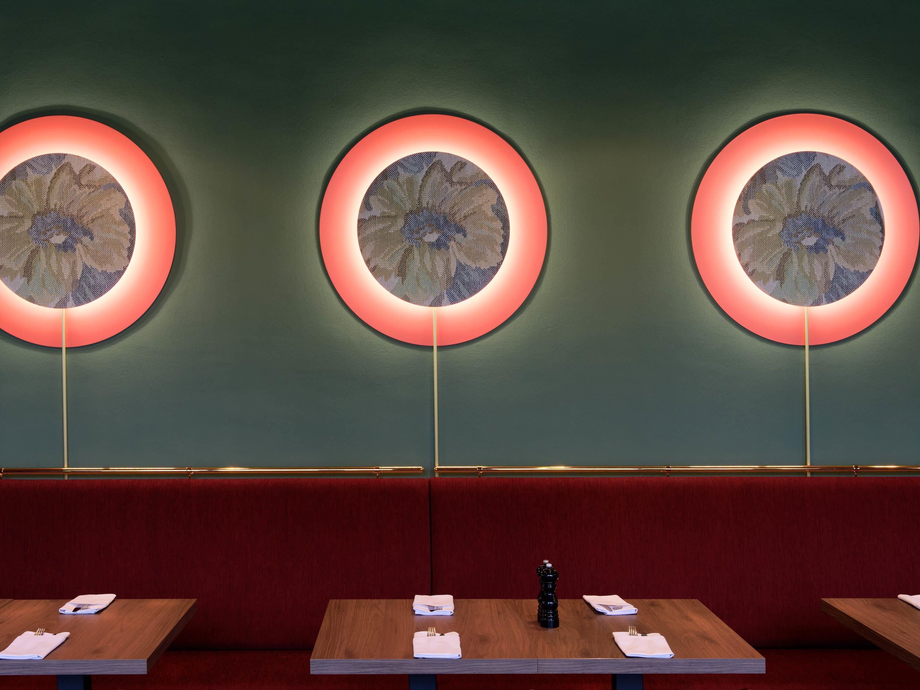 A restaurant interior with three circular wall decorations, wooden tables with folded napkins, and red cushioned seating.