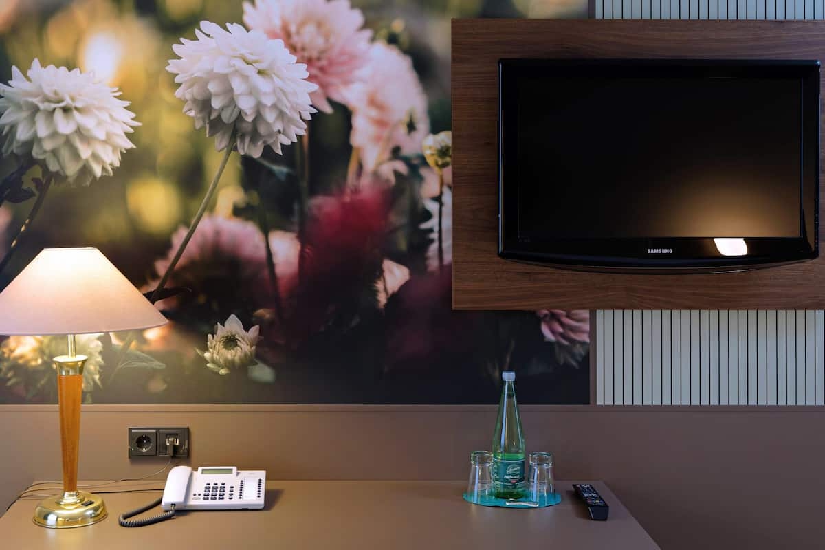 A hotel room with a mounted Samsung TV, a telephone, a lamp, and a minibar with a bottle and glasses.