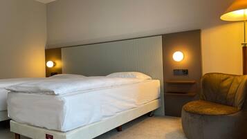 A hotel room with two single beds, a brown armchair, a bedside table with a lamp, and a wall-mounted light.