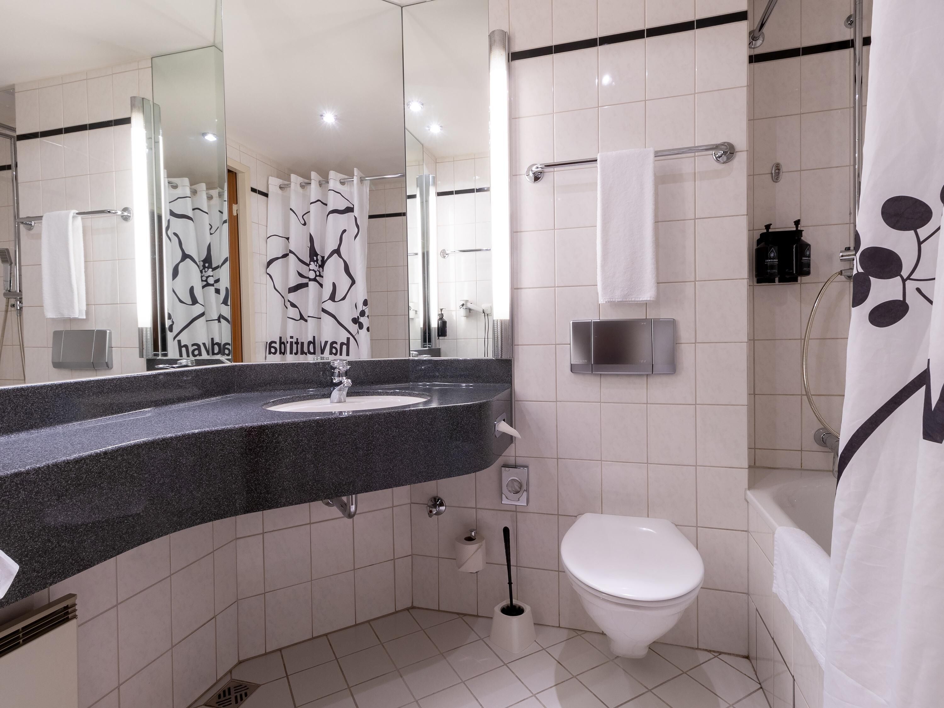 Un baño moderno con lavamanos de granito, inodoro blanco, ducha con puerta de vidrio y espejo con cortina floral.