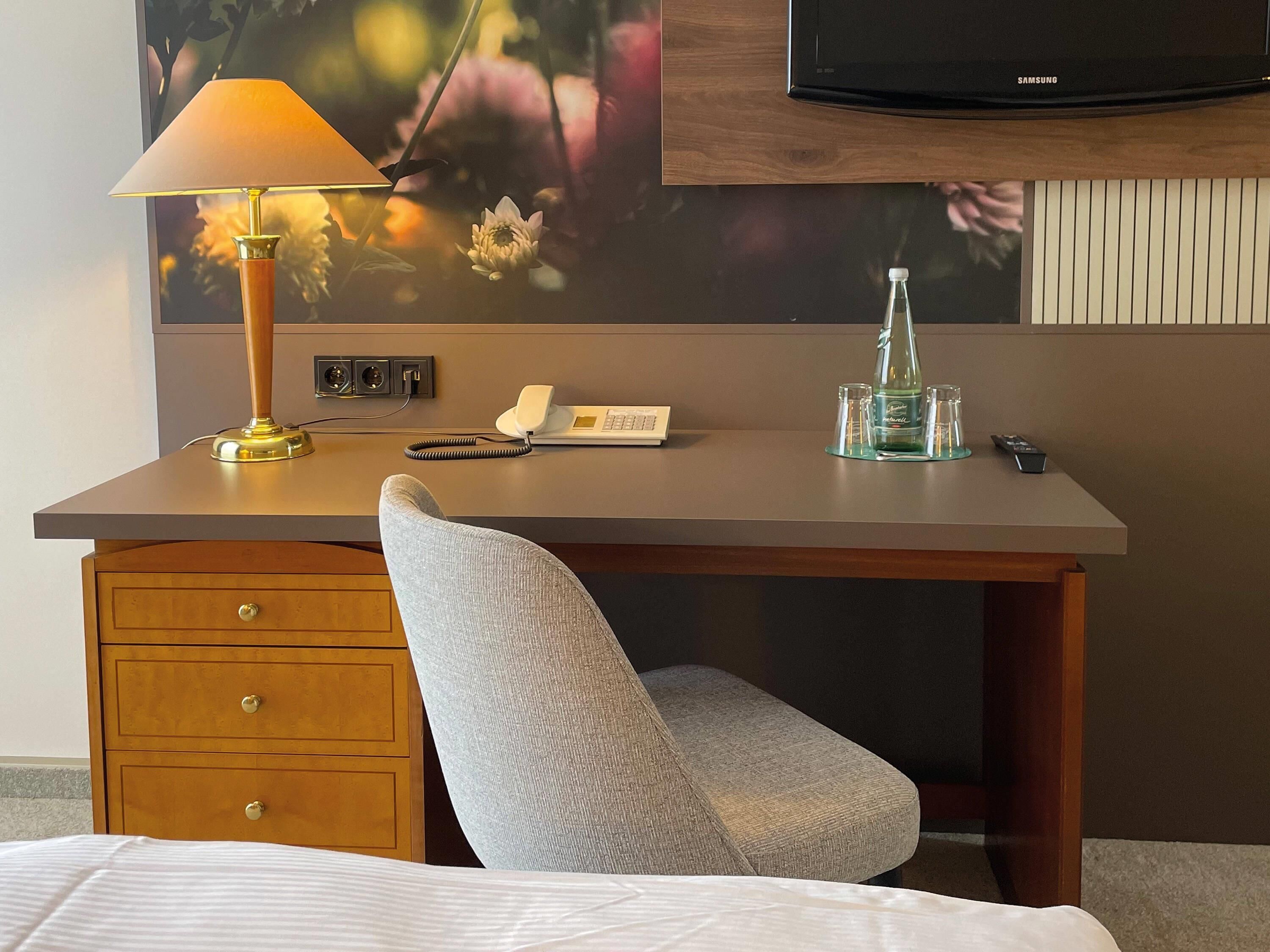 Una habitación de hotel con un escritorio de madera, una lámpara, un teléfono, una botella y un televisor.