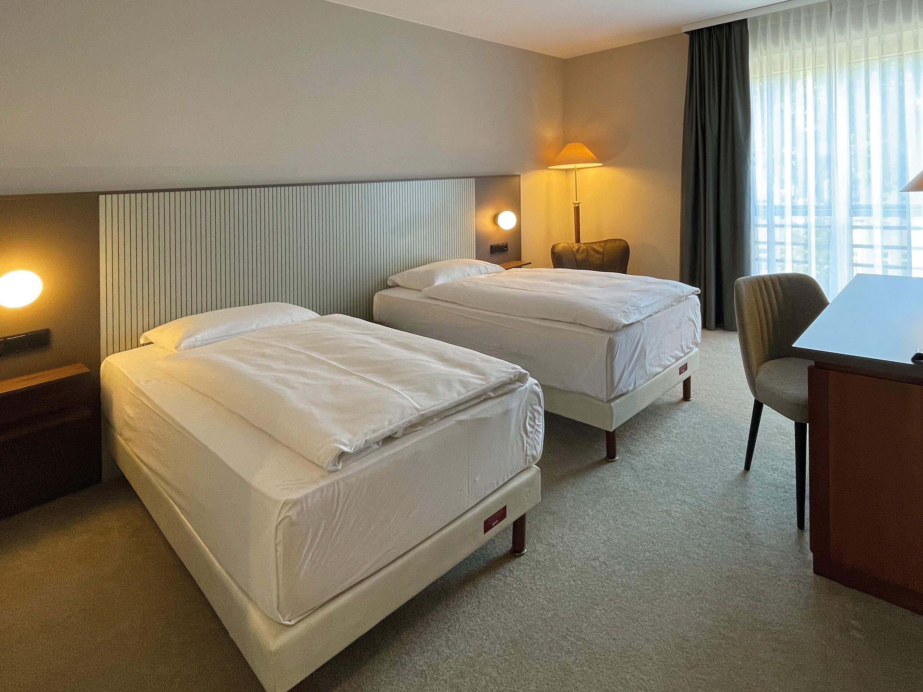 Ein Hotelzimmer mit zwei Betten, einem Schreibtisch, einem Stuhl, einer Lampe und einem Fenster mit Vorhängen.