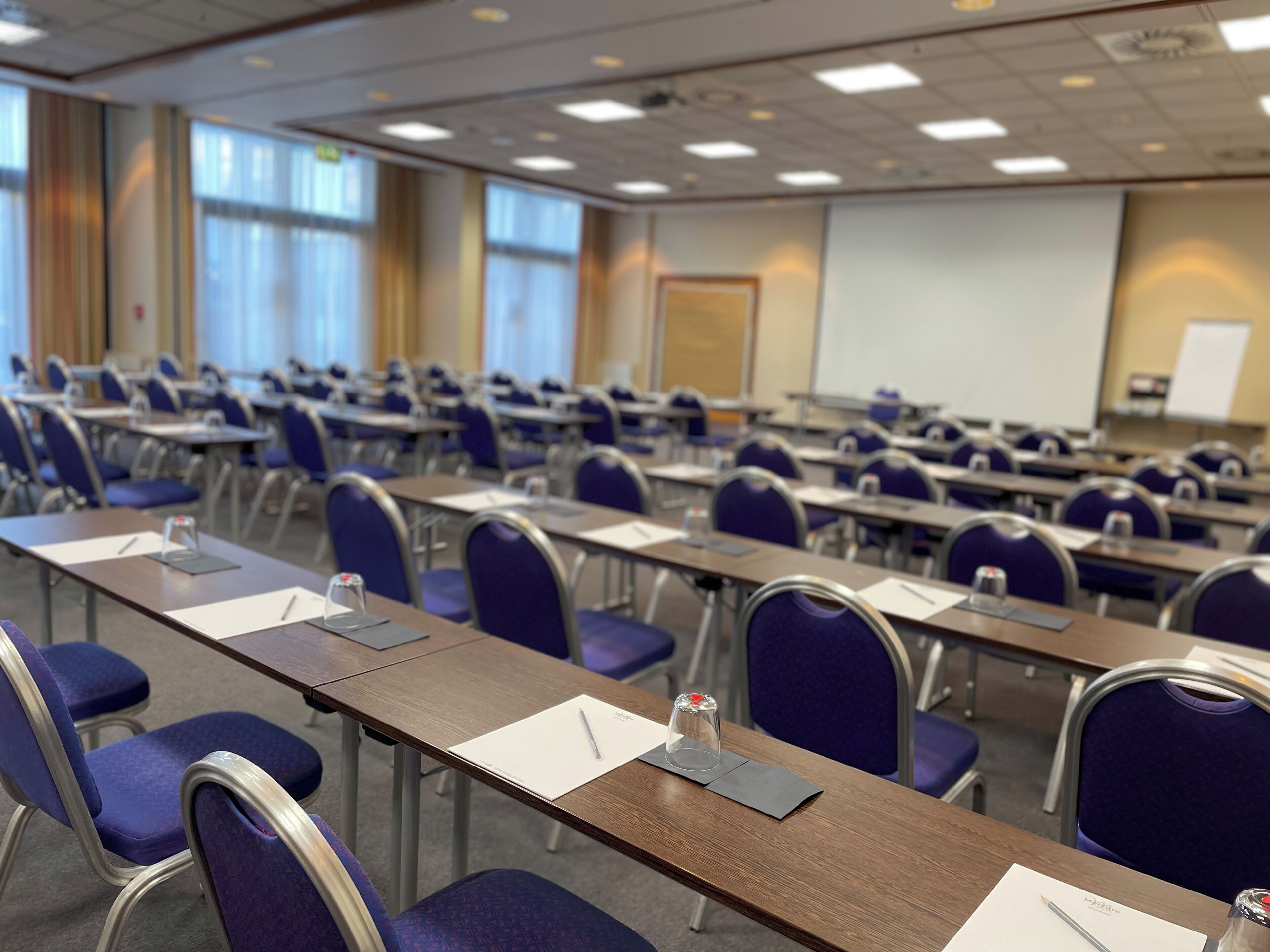 A conference room with rows of tables and chairs, a projector screen, and a whiteboard.
