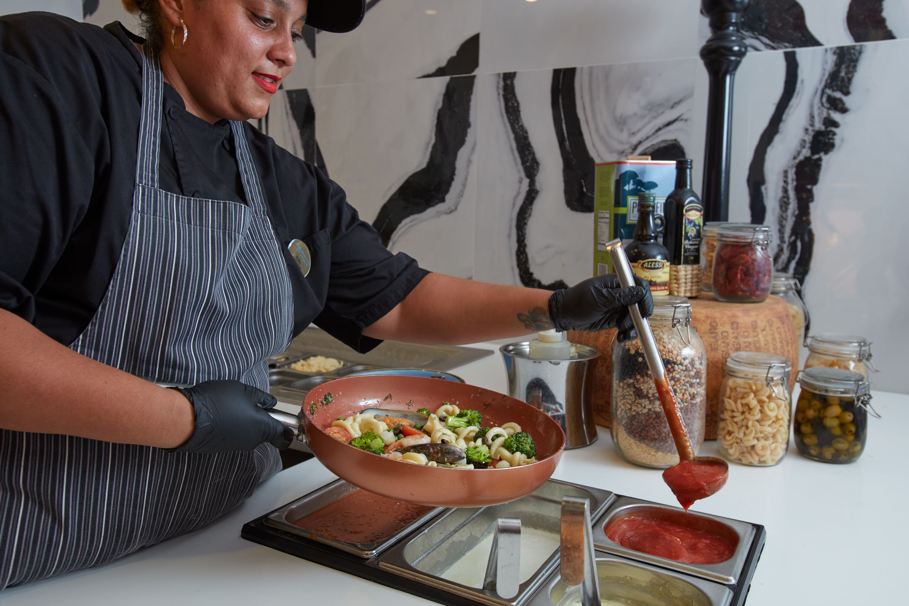 Una persona cocina con una sartén sobre la hornilla, rodeada de diversos ingredientes y condimentos.