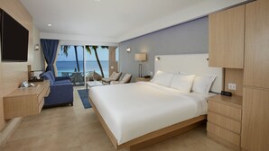 Ein Hotelzimmer mit einem großen Bett, einem Schreibtisch mit einem Laptop, einem Stuhl, einem kleinen Tisch mit einem Glas Wasser und Blick auf den Ozean.