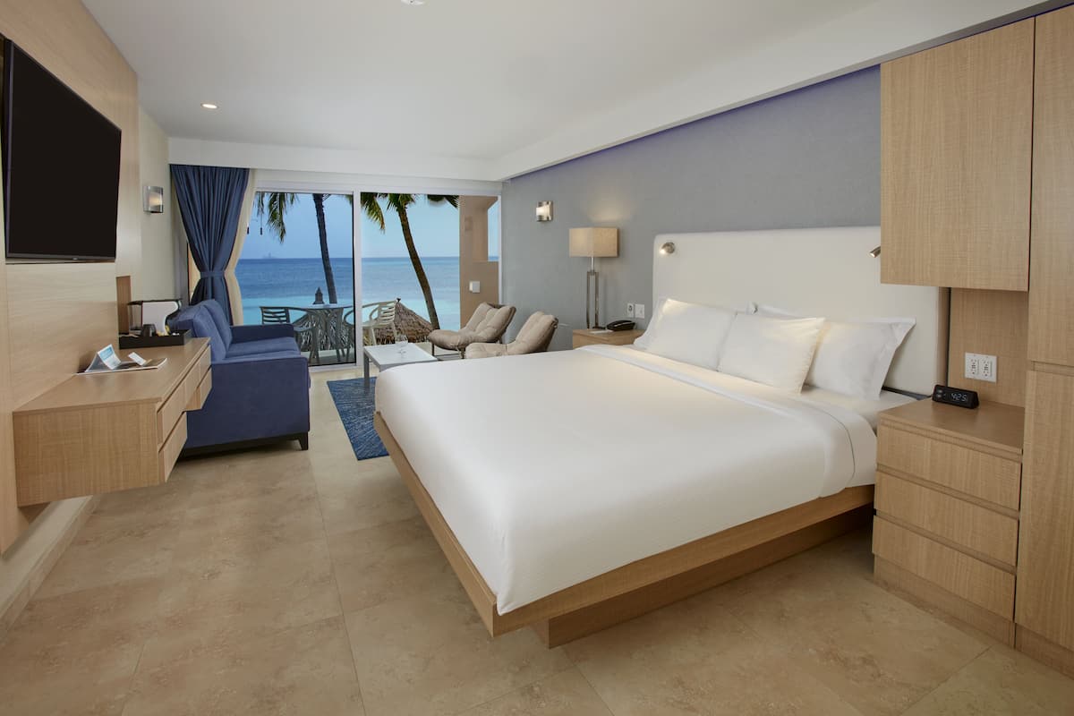 Ein Hotelzimmer mit einem großen Bett, einem Schreibtisch mit einem Laptop, einem Stuhl, einem kleinen Tisch mit einem Glas Wasser und Blick auf den Ozean.