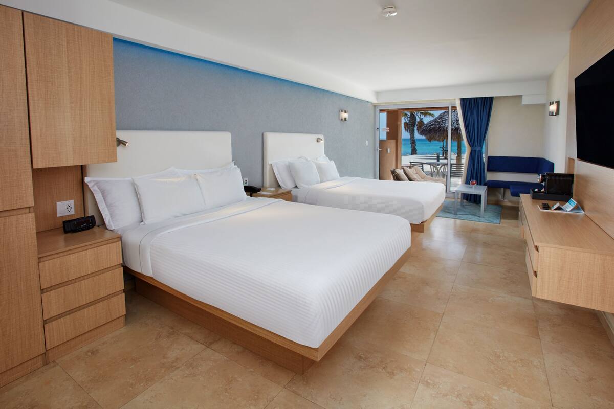 Ein Hotelzimmer mit zwei Betten, einem Schreibtisch, einem Fernseher und Blick auf den Strand.