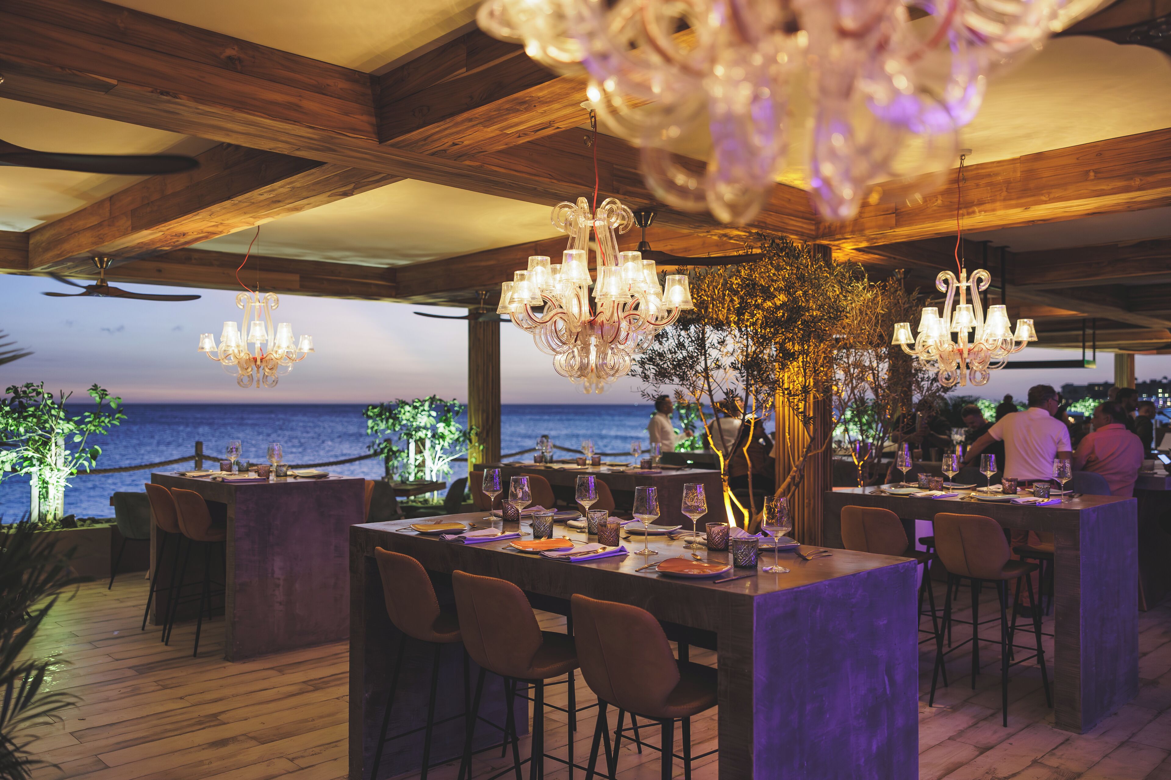 An outdoor dining area with wooden beams, chandeliers, and a view of the sea.