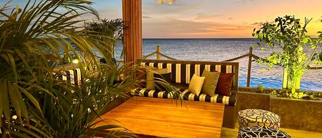 A wooden deck with a sofa and ottomans, a chandelier, and a view of the sea.