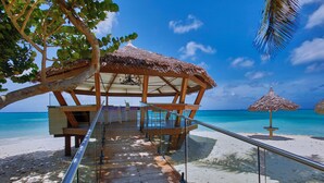 Eine Holzterrasse führt zu einem Gebäude mit einem Strohdach und einem Pool, mit Blick auf einen Strand.