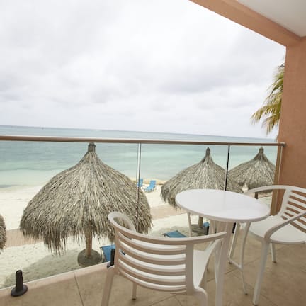 A balcony with a view of the beach, white plastic chairs, and thatched umbrellas.