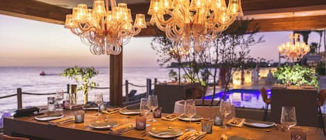 A dining table set for a meal with a view of the sea, elegant chandeliers, and a wooden ceiling.
