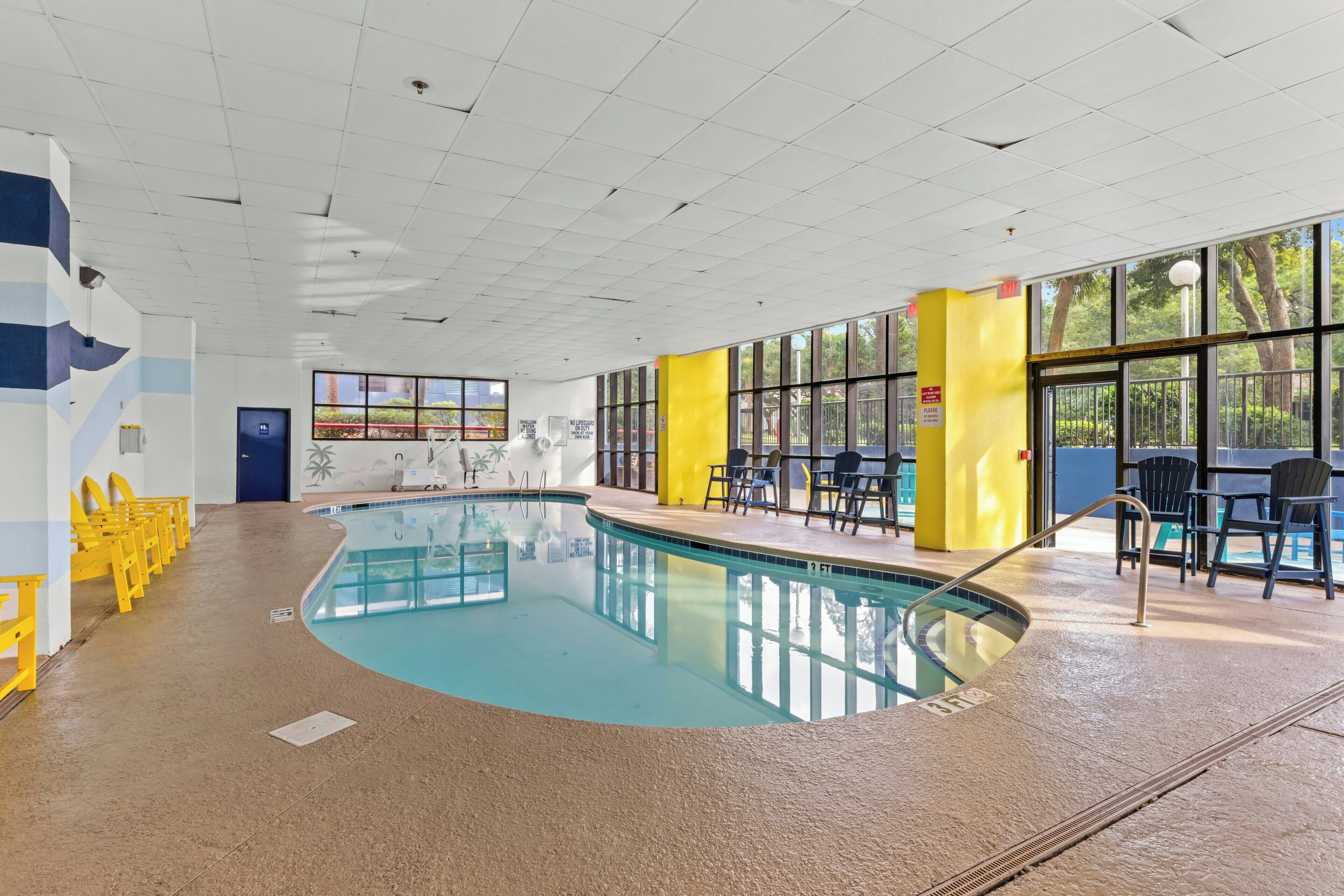 Une piscine intérieure à l’eau claire, avec des chaises jaunes et de grandes fenêtres donnant sur l’extérieur.