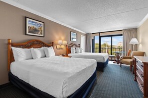 Two large beds with white linens and wooden headboards, a small table, and a chair, set against a backdrop of a large window with an outside view.